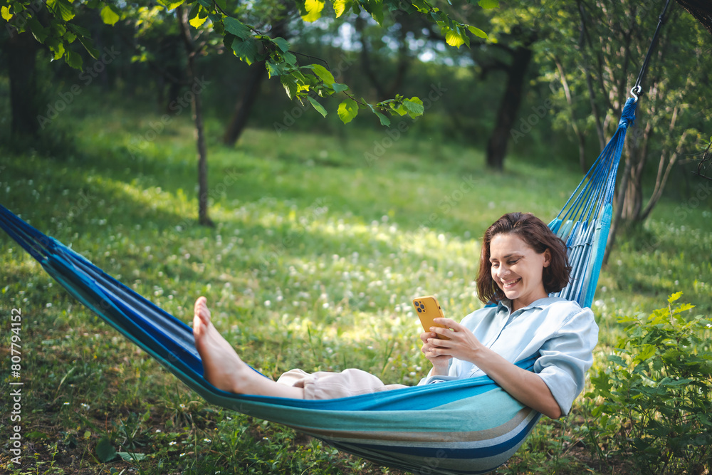 Cheerful young Caucasian woman in a hammock in a summer garden using a smartphone