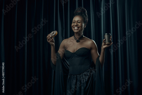 Smiling African-American woman, dressed in vintage black dress holds sweet donut and cup of coffee against dark curtain backdrop. Concept of comparison of eras, modernity and history.