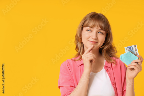 Thoughtful young woman holding money and blue purse on yellow background