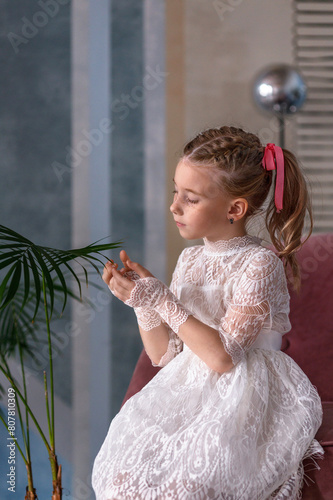 A cute little toddler girl in a white holiday dress sits near a palm tree. Childhood