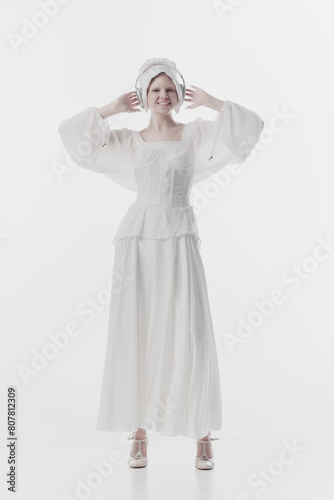 Young lady, in guise of simple peasant woman wearing old fashion clothes listening music in headphones against white studio background. Concept of history, renaissance art, comparison of eras, vintage