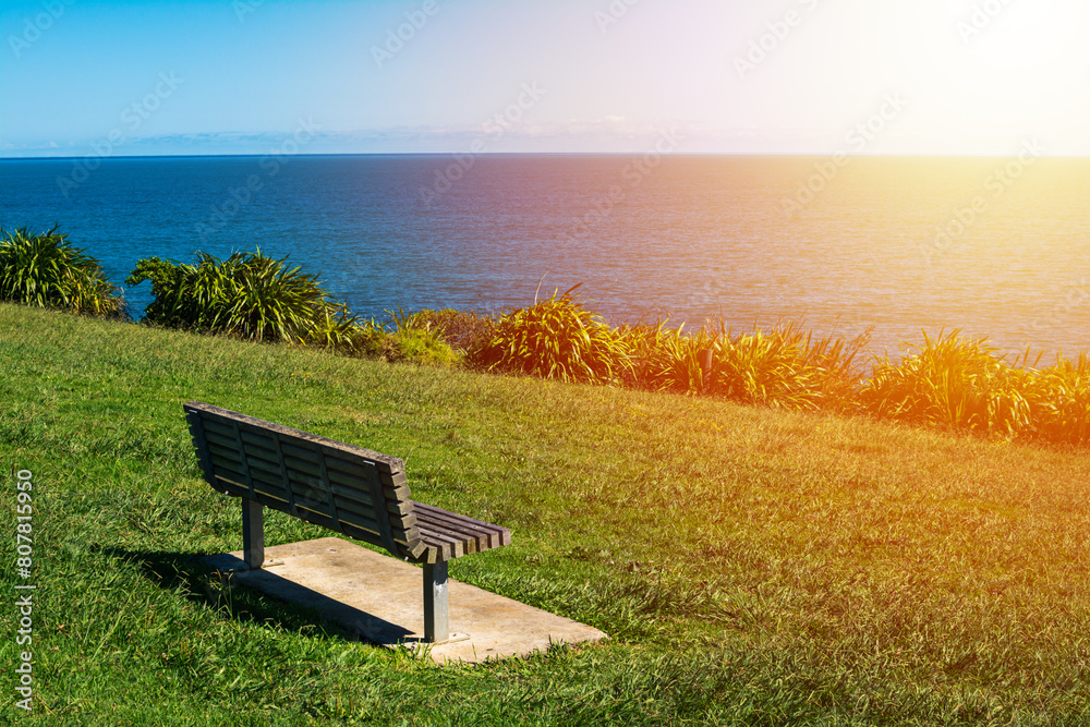 A minimalistic landscape with a bench on green grass looking out toward the ocean sea waters. Blue sky and low clouds over distant mountains. Raglan, New Zealand A minimalistic landscape with a bench on green grass looking out toward the ocean sea waters. Blue sky and low clouds over distant mountains. Raglan, New Zealand