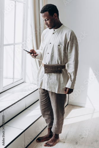Stylishly dressed African American man stands by large window, absorbed in using his smartphone, in spacious and well-lit modern home setting. Clothes made of natural linen fabric, eco-friendly shirt.
