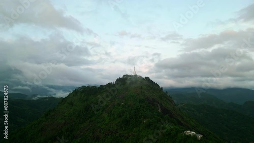 Drone circles Ambuluwawa Tower, rising above mist-shrouded Sri Lankan mountains. Verdant slopes, early morning gilds serene landscape. Sacred architecture, natural wonders attract tourists, hikers. photo