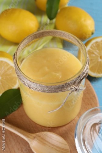 Delicious lemon curd in glass jar, fresh citrus fruits, spoon and green leaves on table, closeup