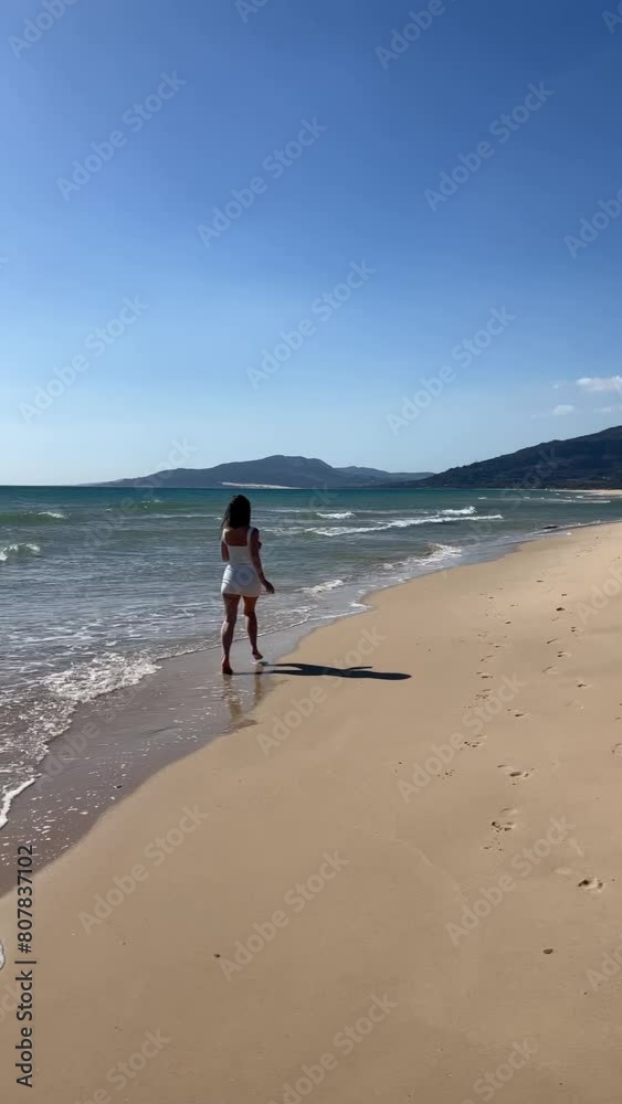 An elegant tourist woman in a white summer dress walks along a tropical beach. An elegant tourist woman in a white summer dress walks along a tropical beach.