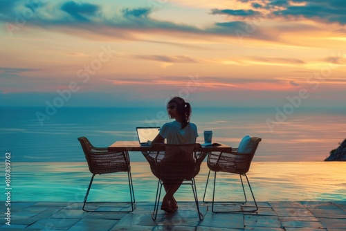 A woman is sitting at a table by the ocean, working on her laptop
