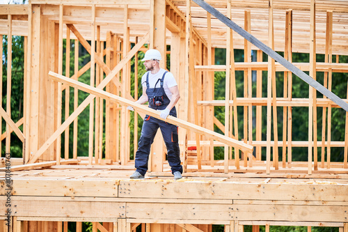 Carpenter constructs wooden-framed house. Bearded man holds large truss in his hands while dressed in work clothes and helmet. An idea of modern and eco-friendly construction.