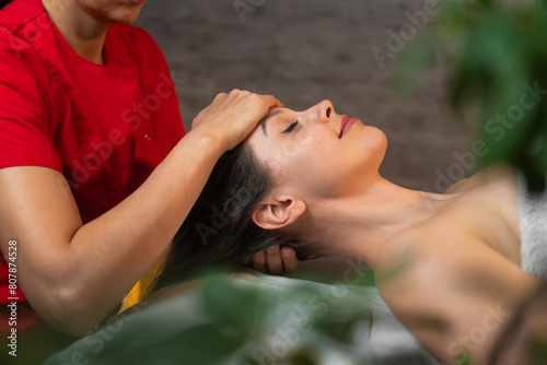 Masseur in a red uniform suit gives a head massage to a young brunette woman lying on a massage table in the spa solon. Spa concept, self-care photo