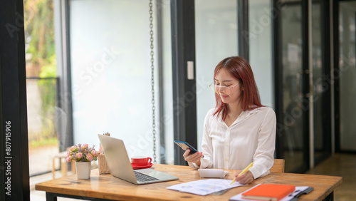 A woman is sitting at a desk with a laptop and a cell phone. She is wearing glasses and has a red phone on her lap. The scene suggests that she is working or studying © apichat