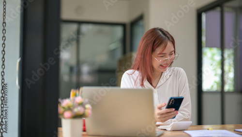 A woman is sitting at a desk with a laptop and a cell phone. She is smiling and she is happy