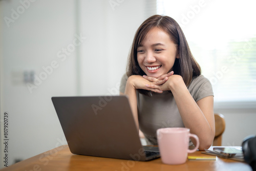 A woman is sitting at a table with a laptop and a pink mug. She is smiling and looking at the camera