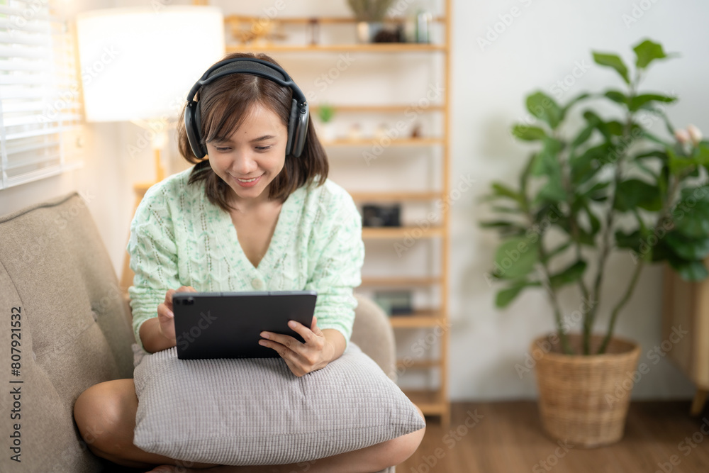 © Wasana - A woman is sitting on a couch with a tablet in her lap. She is wearing headphones and she is enjoying her time. The room has a cozy and comfortable atmosphere, with a potted plant