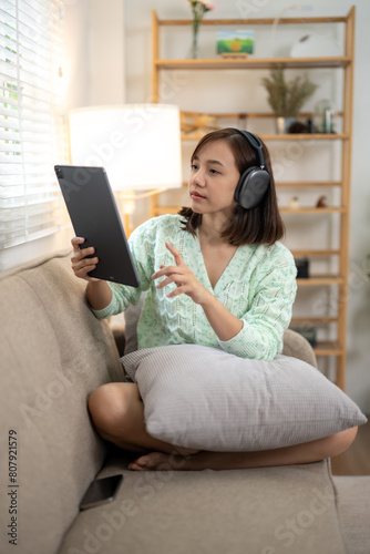 A woman is sitting on a couch with a tablet in her lap. She is wearing headphones and she is engaged in a conversation or activity on the tablet. Concept of relaxation and leisure