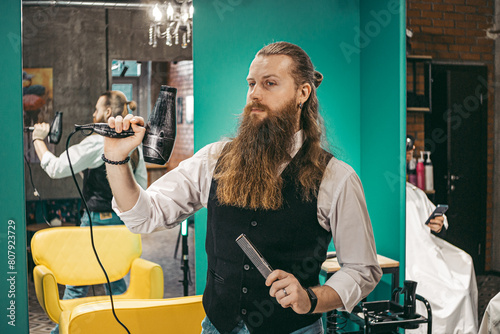 Hairdresser funny shoots a hairdryer. Man young barber barber cheerful joking with long beard and hair in vest  joking shooting with hair dryer laughing in barbershop hairdresser beauty salon.