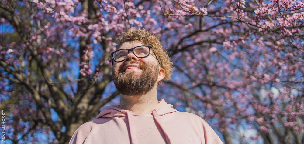 Fototapeta premium Banner Man allergic enjoying after treatment from seasonal allergy at spring. Portrait of happy bearded man smiling in front of blossom tree at springtime. Spring blooming and allergy concept. Copy