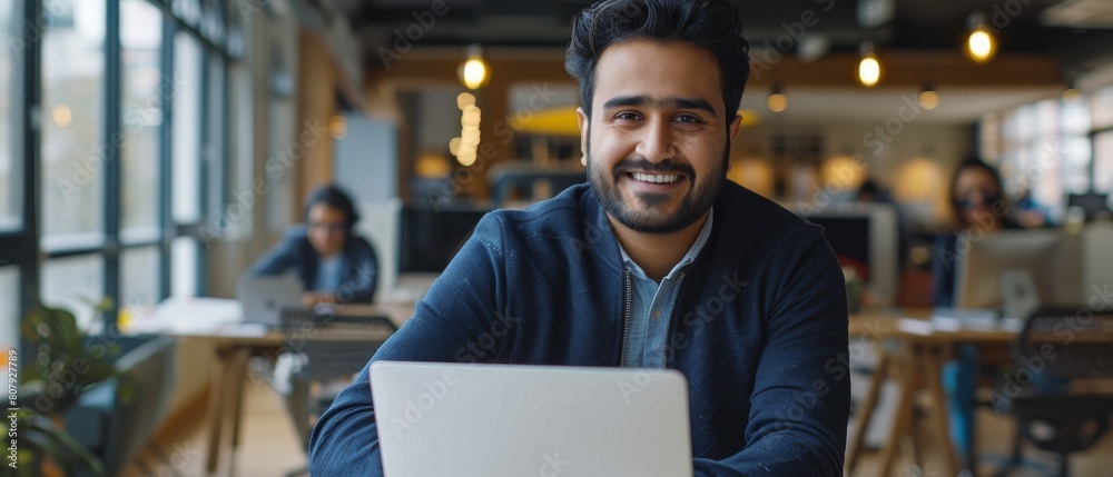 Working on a laptop, a handsome, smiling Indian office worker sits at his desk. In the background, a modern office filled with young professionals works. Working on a laptop, a handsome, smiling Indian office worker sits at his desk. In the background, a modern office filled with young professionals works.