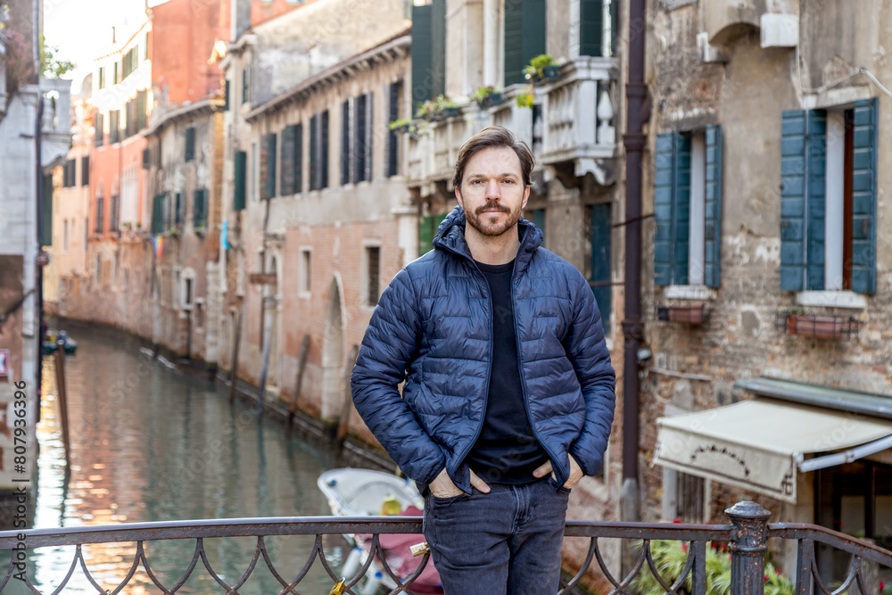 Obraz premium Bearded man wearing casual clothes stands leaning against railings on a bridge in Venice; Venetian canal in the background