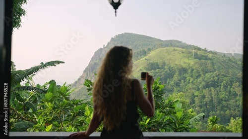 Blonde woman sips coffee at upscale resort balcony with breathtaking Ella Rock mountain landscape in backdrop. Leisurely enjoying morning amidst Sri Lankan green plants, embodies serene travel luxury.