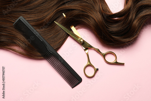 Professional hairdresser scissors and comb with brown hair strand on pink background, top view