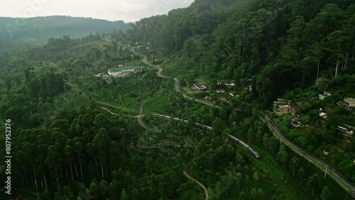 Aerial view of train journey through Haputale mountain landscapes passes tea plantations, misty forests in Sri Lanka. Clouds hover as locomotive snakes along tracks, surrounded by lush green plants. photo