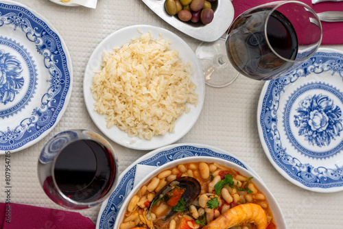 A dish of seafood feijoada with rice, garnished with shrimp and clams, sits on a restaurant table. Alongside are Alentejo cheese and a glass of red wine, evoking a warm Portuguese dining experience. photo