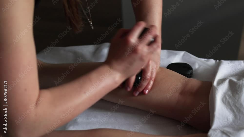 Woman enjoying hot stone massage on her legs at spa salon. Professional masseur making stone therapy. Relaxing and ease tense muscles and damaged soft tissues throughout body. Woman enjoying hot stone massage on her legs at spa salon. Professional masseur making stone therapy. Relaxing and ease tense muscles and damaged soft tissues throughout body.
