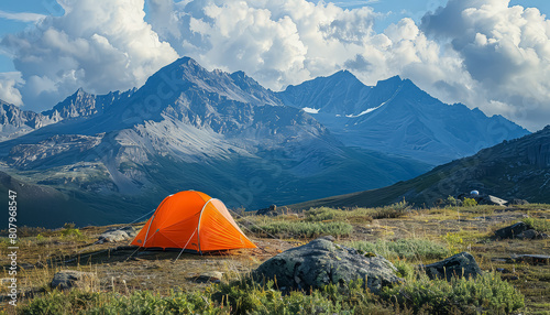 A small orange tent is set up on a grassy hillside in front of a mountain range