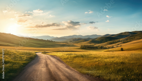 A dirt road winds through a grassy field at sunset leads to a mountain range in the distance with blue sky and clouds.