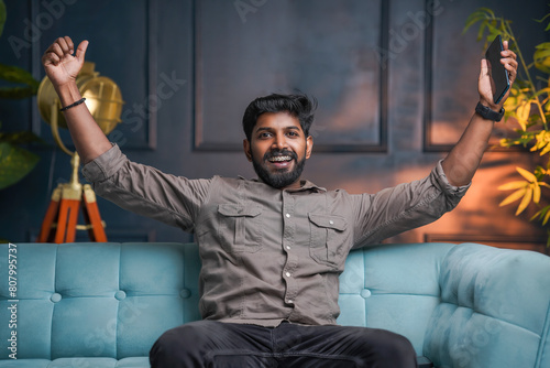 Excited Indian man sitting on sofa and rising his hand with mobile phone © sarath