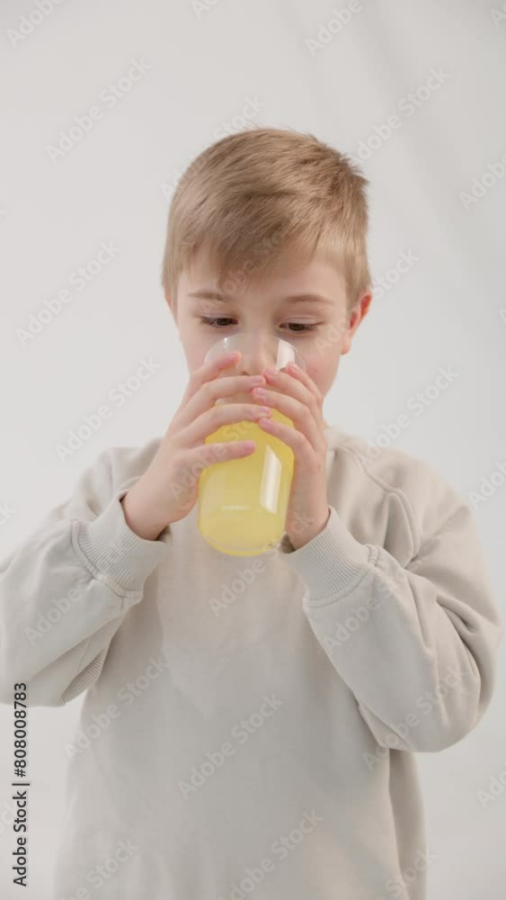 Vertical video. Caucasian blond boy with blue eyes drinks lemonade from a glass and starts licking himself. White background behind. The concept of healthy natural food. Vertical video. Caucasian blond boy with blue eyes drinks lemonade from a glass and starts licking himself. White background behind. The concept of healthy natural food.