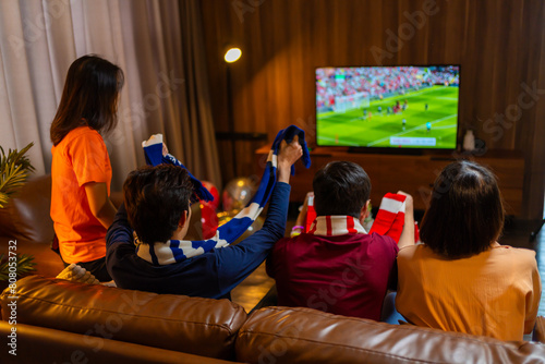 Group of Asian people friends sit on sofa watching and cheering football or soccer games competition on TV together at home.Happy man and woman sport fans celebrating sport team victory sports match © Yingyaipumi