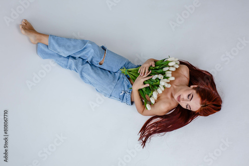 Fototapeta Naklejka Na Ścianę i Meble -  Red-haired girl in jeans with white tulips posing while lying on a white background