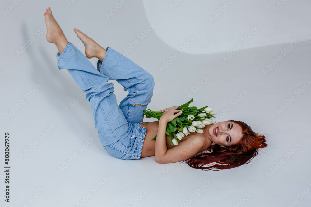 Red-haired girl in jeans with white tulips posing while lying on a white background
