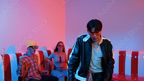 Asian man standing and dancing with pop music while multicultural friend sitting behind at home with led light. Attractive street dancer moving to hip hop music while looking at camera. Regalement. photo
