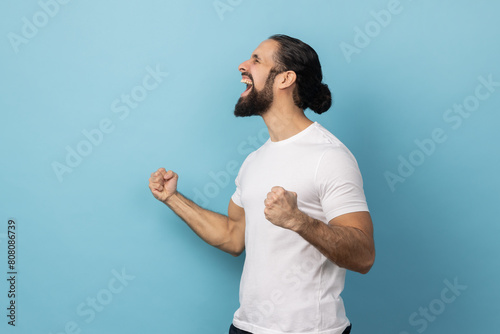 Side view of excited man with beard wearing white T-shirt expressing winning gesture with raised fists and screaming, celebrating victory. Indoor studio shot isolated on blue background.