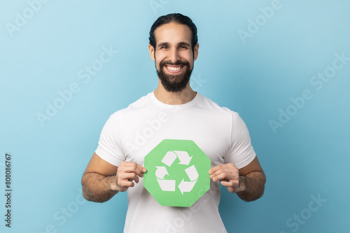 Portrait of smiling man with beard wearing white T-shirt showing green waste recycling symbol, satisfied with environmental safety. Indoor studio shot isolated on blue background.