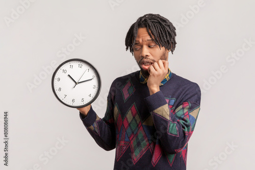 Portrait of nervous sad upset african-american man with dreadlocks and beard holding big wall clock, biting fingernails, being late. Indoor studio shot isolated on gray background.