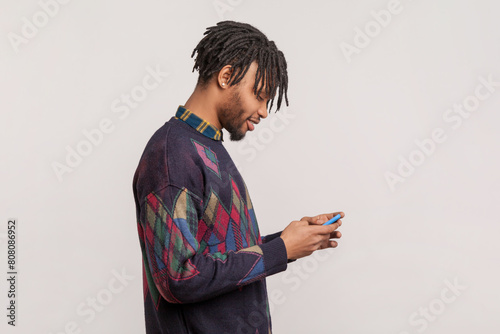 Side view portrait of smiling happy african-american man with dreadlocks and beard standing with mobile phone in hands, checking social networks. Indoor studio shot isolated on gray background.