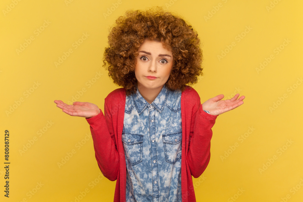 Portrait of puzzled clueless woman with Afro hairstyle receives strange offer, shrugging shoulders with hesitation, feels doubt. Indoor studio shot isolated on yellow background.
