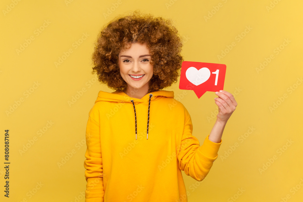 Cheerful carefree woman with Afro hairstyle holding Heart Like icon of social media, notification button to follow, subscribe blog, popular forum. Indoor studio shot isolated on yellow background.