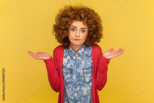 Portrait of puzzled clueless woman with Afro hairstyle receives strange offer, shrugging shoulders with hesitation, feels doubt. Indoor studio shot isolated on yellow background.