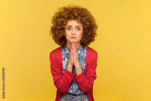 Portrait of pleading woman with Afro hairstyle holding hands folded in praying gesture, asking for help, pressing palms together. Indoor studio shot isolated on yellow background.
