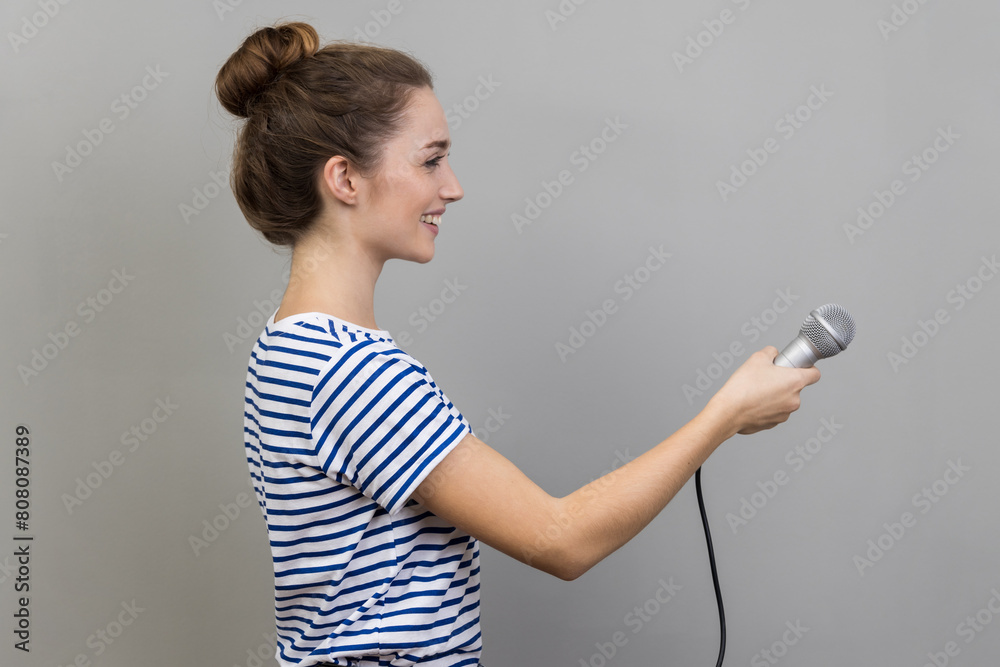 Side view of smiling friendly beautiful woman wearing striped T-shirt standing offering microphone, journalist taking interview. Indoor studio shot isolated on gray background. Side view of smiling friendly beautiful woman wearing striped T-shirt standing offering microphone, journalist taking interview. Indoor studio shot isolated on gray background.