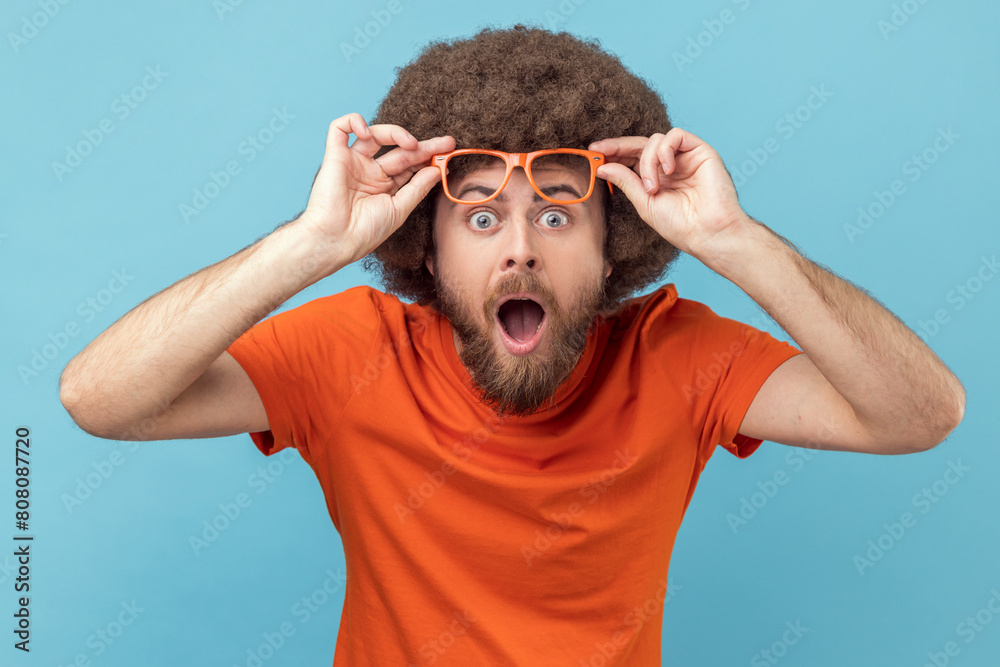 Φωτογραφία Shocked amazed man with Afro hairstyle wearing orange T-shirt standing and looking at camera, raising his eyeglasses, sees something astonishing