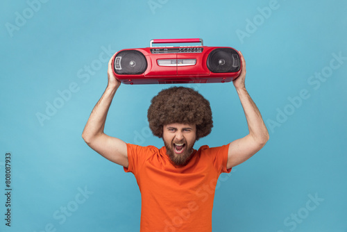 Portrait of crazy man with Afro hairstyle in orange T-shirt listening to music, holding red tape recorder above head, screaming, having fun on party. Indoor studio shot isolated on blue background.