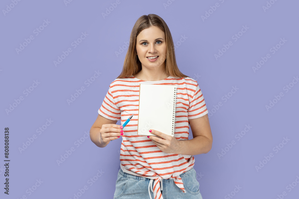 Portrait of attractive optimistic woman in striped T-shirt holding organizer with empty paper with advertisement area, pointing at notebook with pen. Indoor studio shot isolated on purple background. Portrait of attractive optimistic woman in striped T-shirt holding organizer with empty paper with advertisement area, pointing at notebook with pen. Indoor studio shot isolated on purple background.