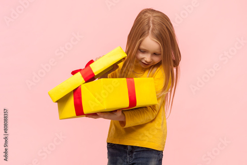 Happy satisfied blonde little girl holding big yellow gift box, looking inside being glad to get present on her birthday. wearing yellow jumper. Indoor studio shot isolated on pink background.