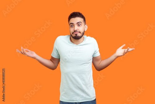 Portrait of uncertain puzzled young bearded man wearing T-shirt shrugging shoulders spreading arms doesn't know answer. Indoor studio shot isolated on orange background.