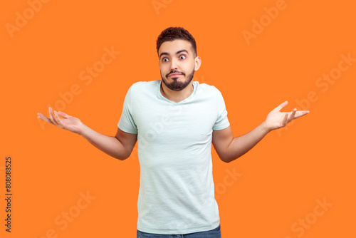 Photography Portrait of uncertain puzzled young bearded man wearing T-shirt shrugging shoulders spreading arms doesn't know answer
